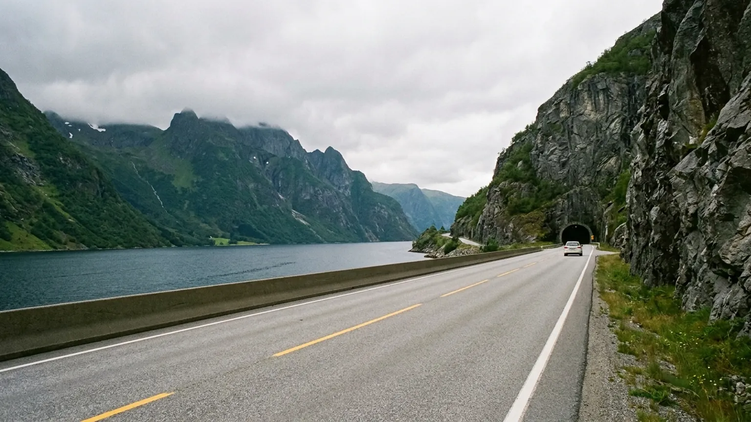 En tofelts vei langs en dyp fjord, over en kort bro og inn i en tunnel i fjellsiden, sett fra høyden.
