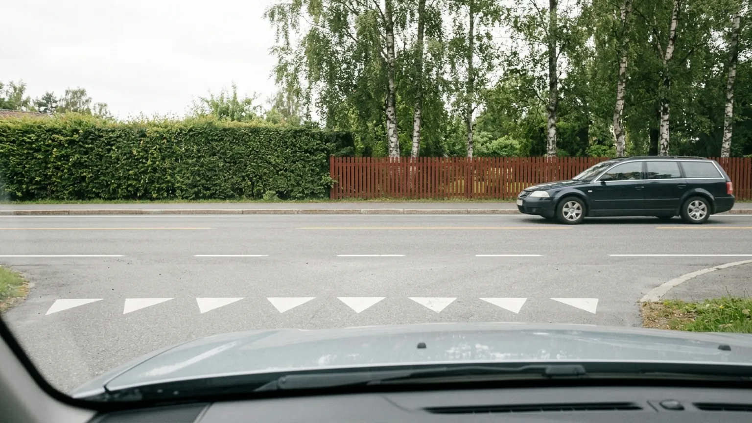 En bil venter i munningen av en sidevei mens en annen bil passerer på hovedveien, sett fra fortauet.