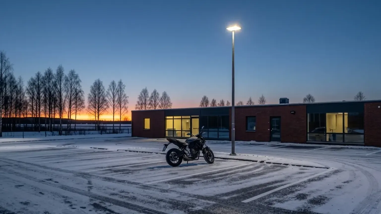 En motorsykkel står alene på en snødekt parkeringsplass foran et lavt murbygg i grålysningen.