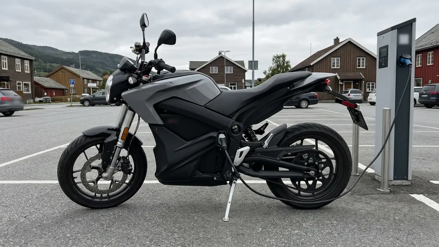 An electric motorcycle stands on its side stand, charging at a post in a car park, wooden houses behind.